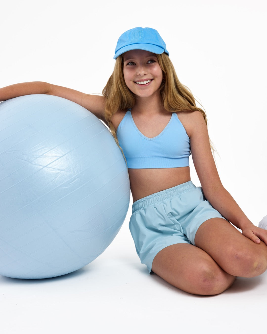 Girl wearing a light blue SASSH Essential training bra, front view, V-neck triangle style with adjustable straps and bow detail — girls first bra.