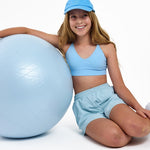 Girl wearing a light blue SASSH Essential training bra, front view, V-neck triangle style with adjustable straps and bow detail — girls first bra.