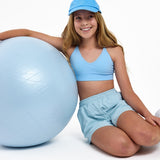 Girl wearing a light blue SASSH Essential training bra, front view, V-neck triangle style with adjustable straps and bow detail — girls first bra.