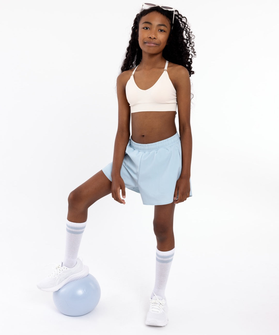 Girl wearing a white SASSH Essential training bra, front view, V-neck triangle style with adjustable straps and bow detail — girls first bra.