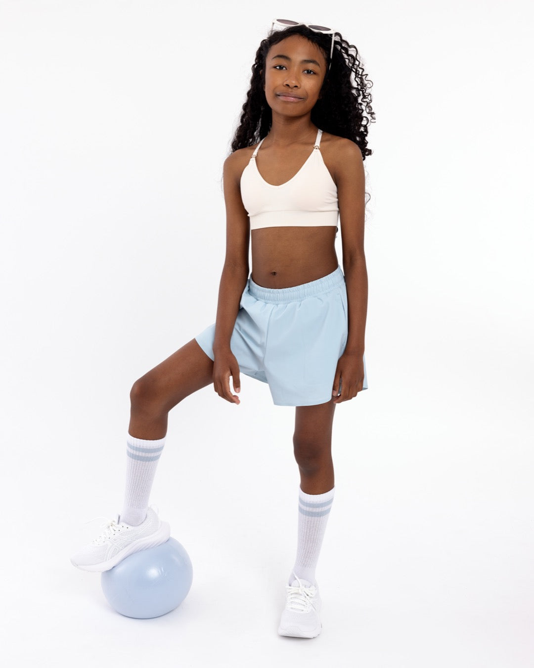 Girl wearing a white SASSH Essential training bra, front view, V-neck triangle style with adjustable straps and bow detail — girls first bra.