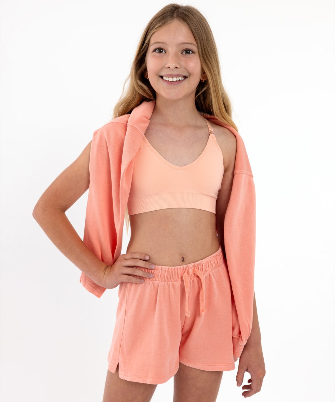 Girl wearing a peach SASSH Essential training bra, front view, V-neck triangle style with adjustable straps and bow detail — girls first bra.