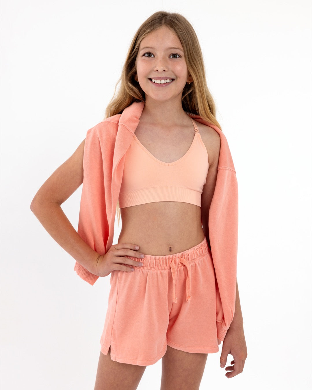 Girl wearing a peach SASSH Essential training bra, front view, V-neck triangle style with adjustable straps and bow detail — girls first bra.