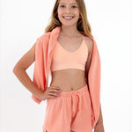 Girl wearing a peach SASSH Essential training bra, front view, V-neck triangle style with adjustable straps and bow detail — girls first bra.