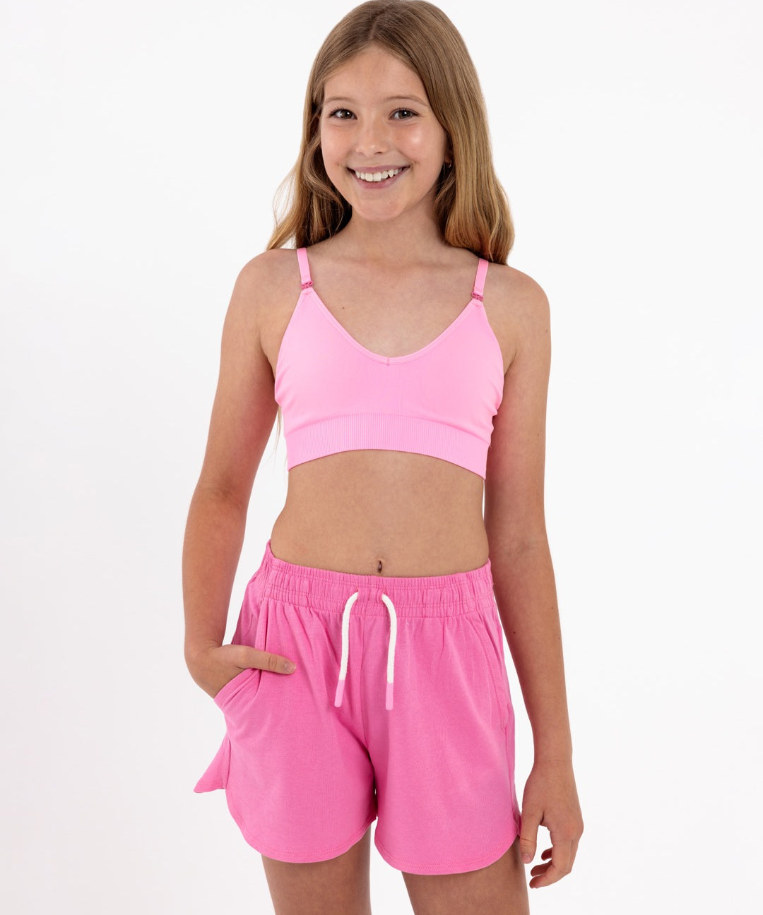 Girl wearing a bright pink SASSH Essential training bra, front view, V-neck triangle style with adjustable straps and bow detail — girls first bra.