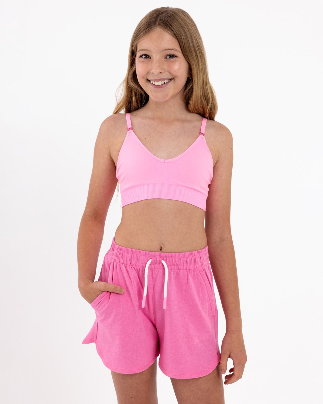 Girl wearing a bright pink SASSH Essential training bra, front view, V-neck triangle style with adjustable straps and bow detail — girls first bra.