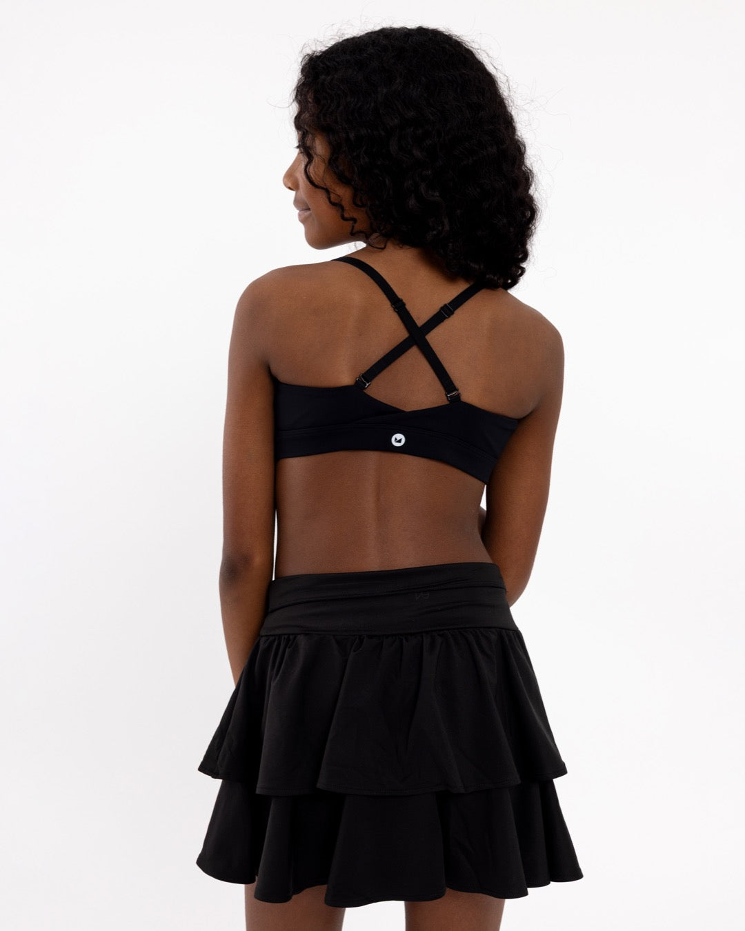 Back view of a girl wearing a black sports bra showing crossover detail, adjustable straps and a pullover style in a white background.