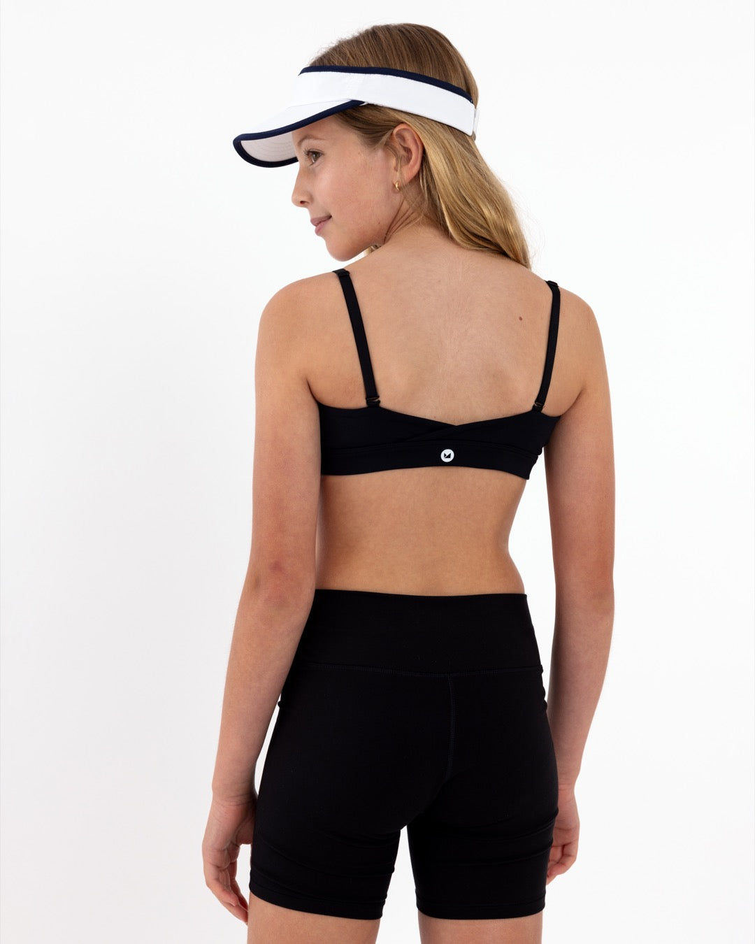 A tween girl wearing a black sports bra and shorts with a white visor on a white background.