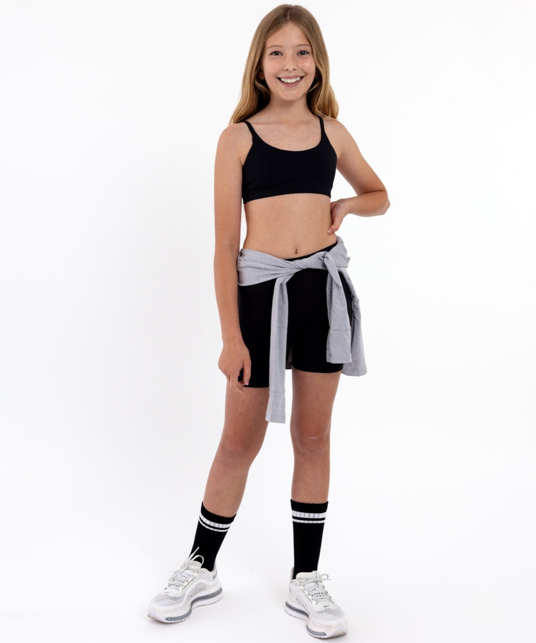 Girl wearing a black sports bra and shorts with a grey jacket wrapping around the waist in a white background.