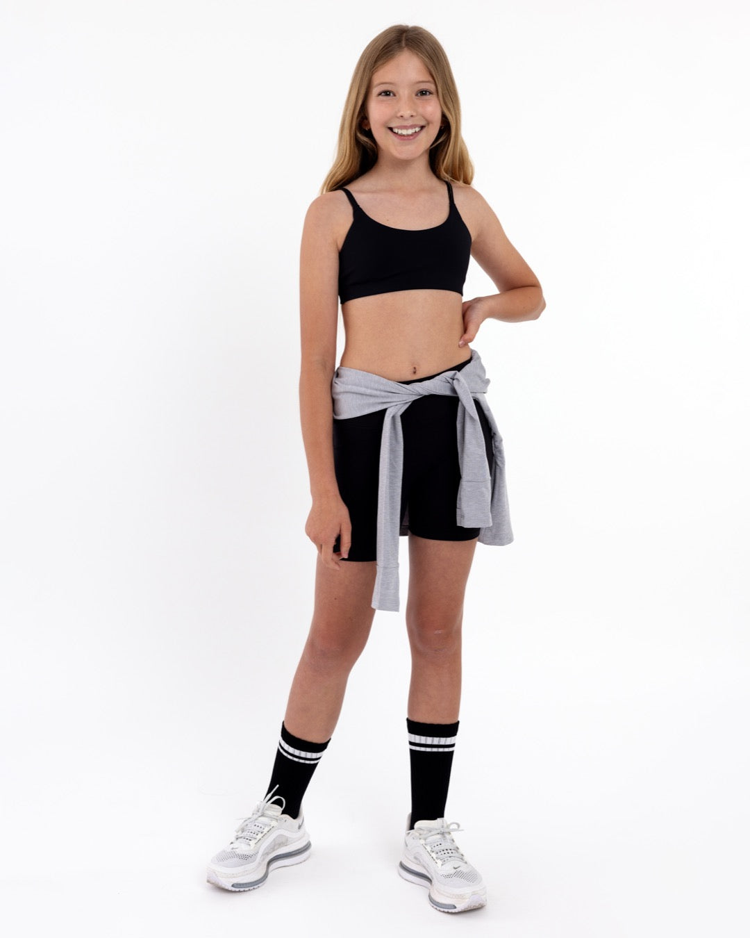 Girl wearing a black sports bra and shorts with a grey jacket wrapping around the waist in a white background.