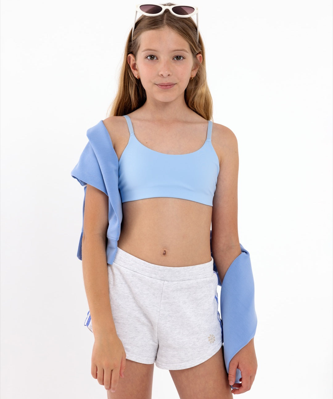 An tween girl in light blue crop top and white shorts with a blue jacket over her shoulder on a white background.