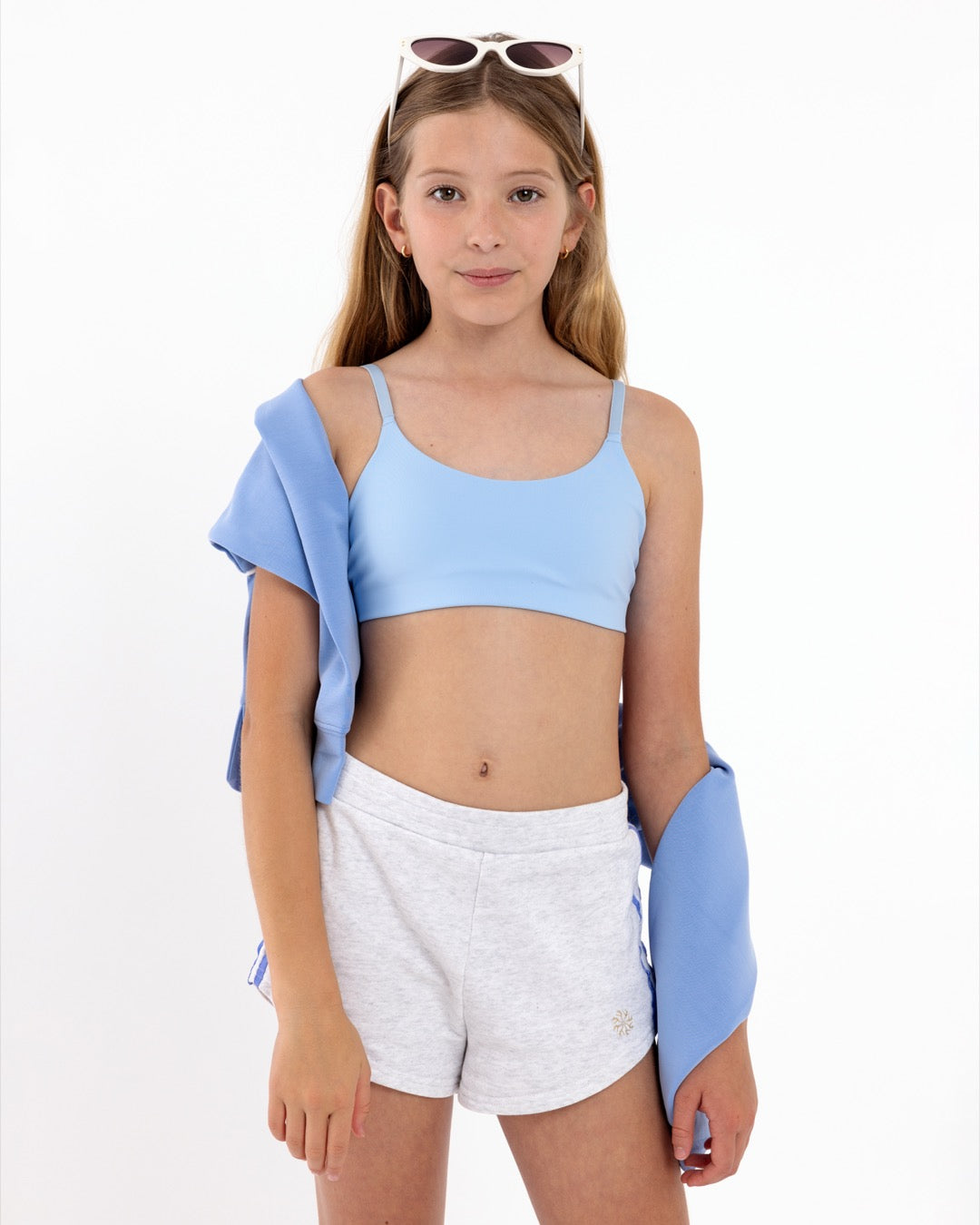 An tween girl in light blue crop top and white shorts with a blue jacket over her shoulder on a white background.