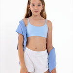An tween girl in light blue crop top and white shorts with a blue jacket over her shoulder on a white background.
