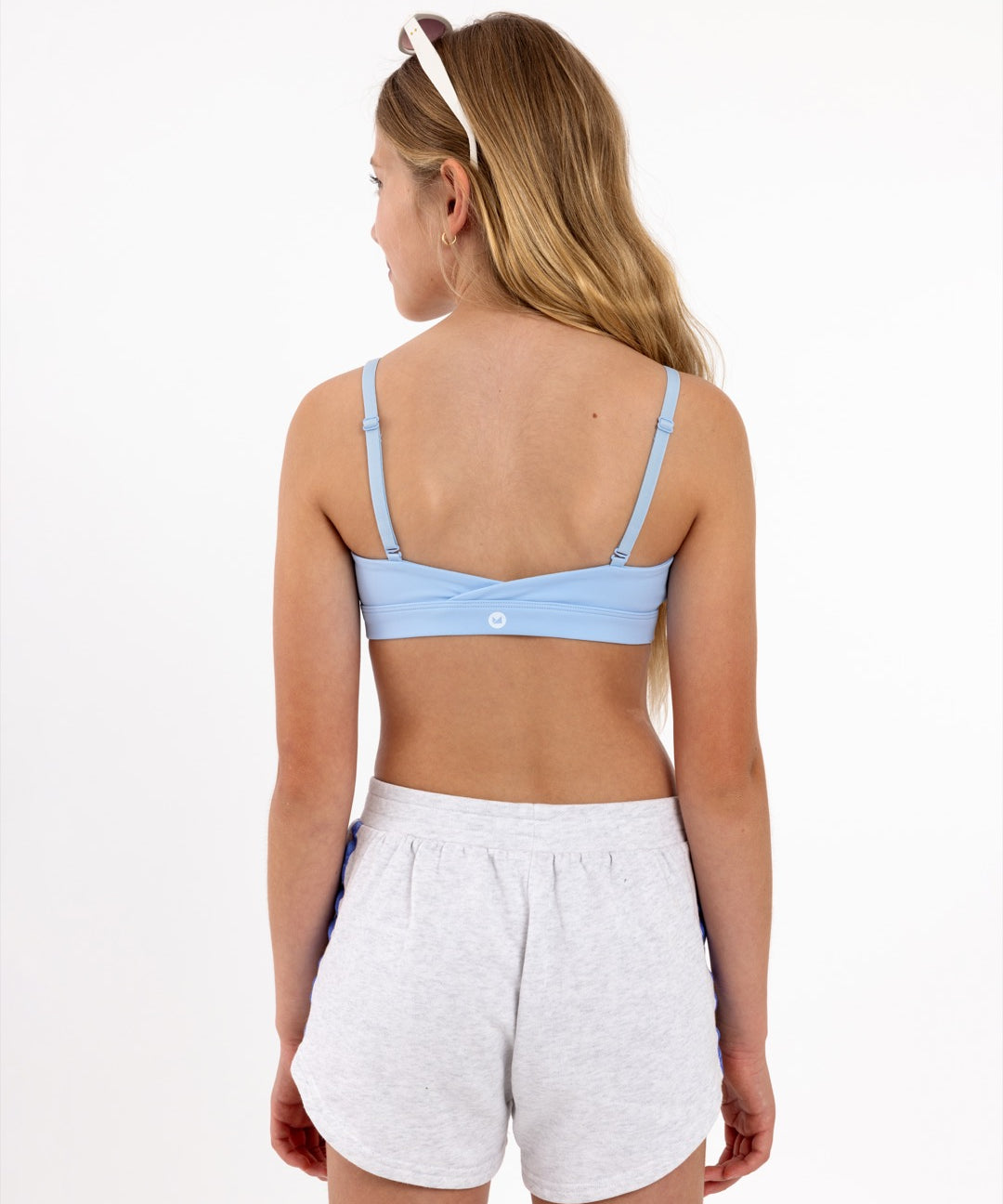 Back view of a girl wearing a light blue sports bra showing crossover detail, adjustable straps and a pullover style in a white background.