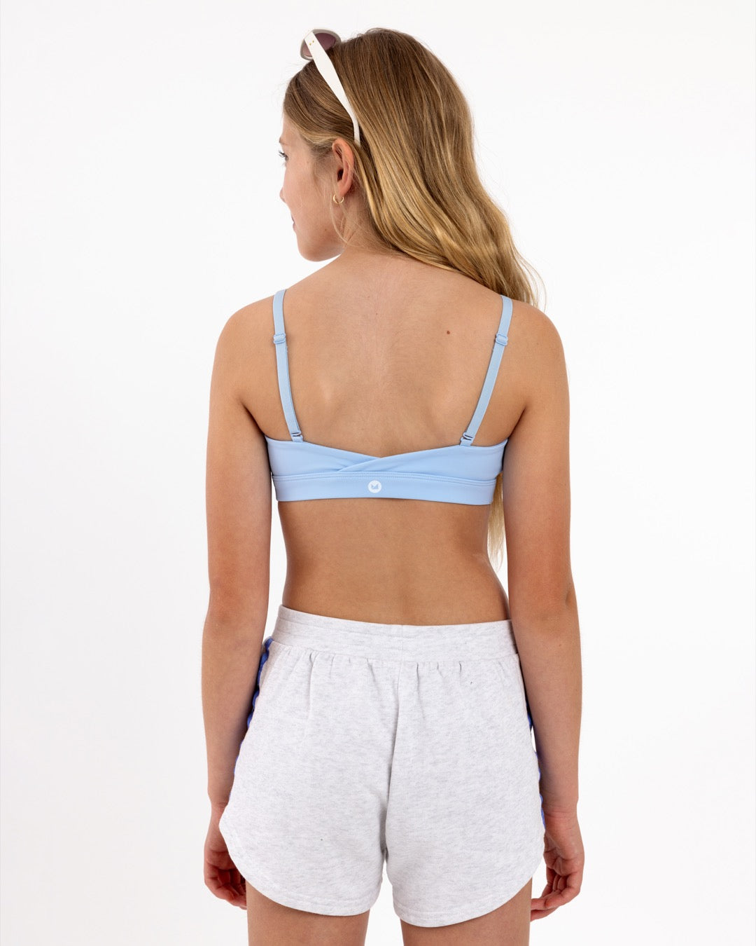 Back view of a girl wearing a light blue sports bra showing crossover detail, adjustable straps and a pullover style in a white background.