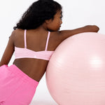 Back view of a girl wearing a pink sports bra leaning on a pink Pilates ball on a white background.