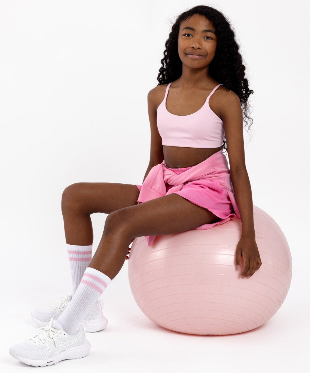 Girl sitting on a pink Pilates ball wearing a pink sports bra with a scoop neckline in a pullover style on a white background.