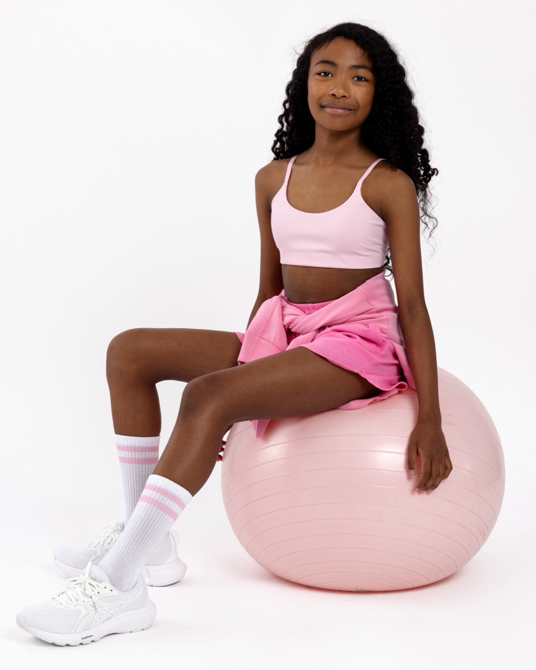 Girl sitting on a pink Pilates ball wearing a pink sports bra with a scoop neckline in a pullover style on a white background.