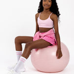 Girl sitting on a pink Pilates ball wearing a pink sports bra with a scoop neckline in a pullover style on a white background.