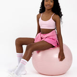 Girl sitting on a pink Pilates ball wearing a pink sports bra with a scoop neckline in a pullover style on a white background.