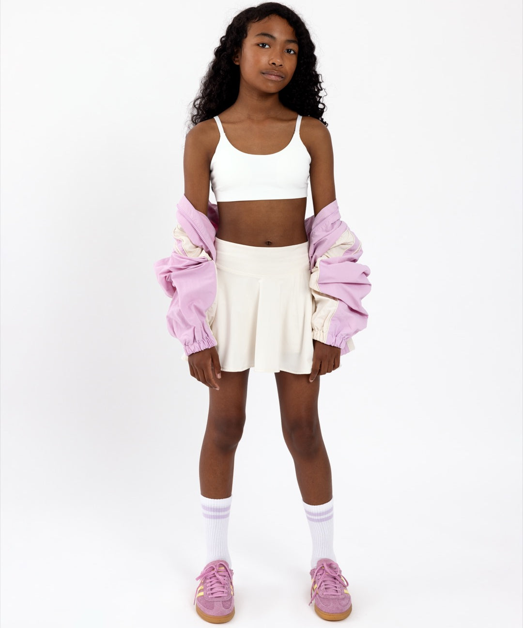 Girl wearing a white sports bra with a scoop neckline, with a white skirt and pink jacket on a white background.