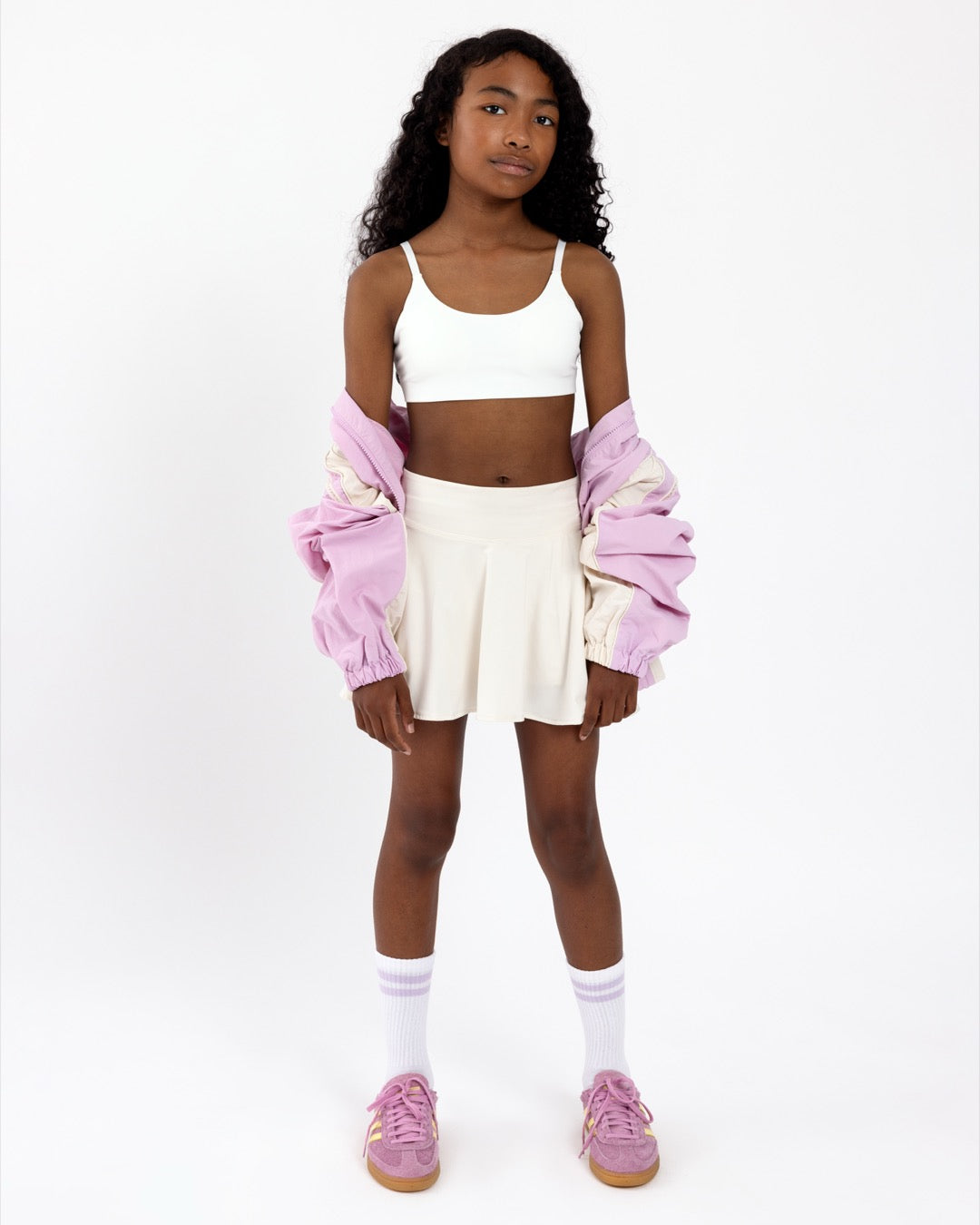Girl wearing a white sports bra with a scoop neckline, with a white skirt and pink jacket on a white background.