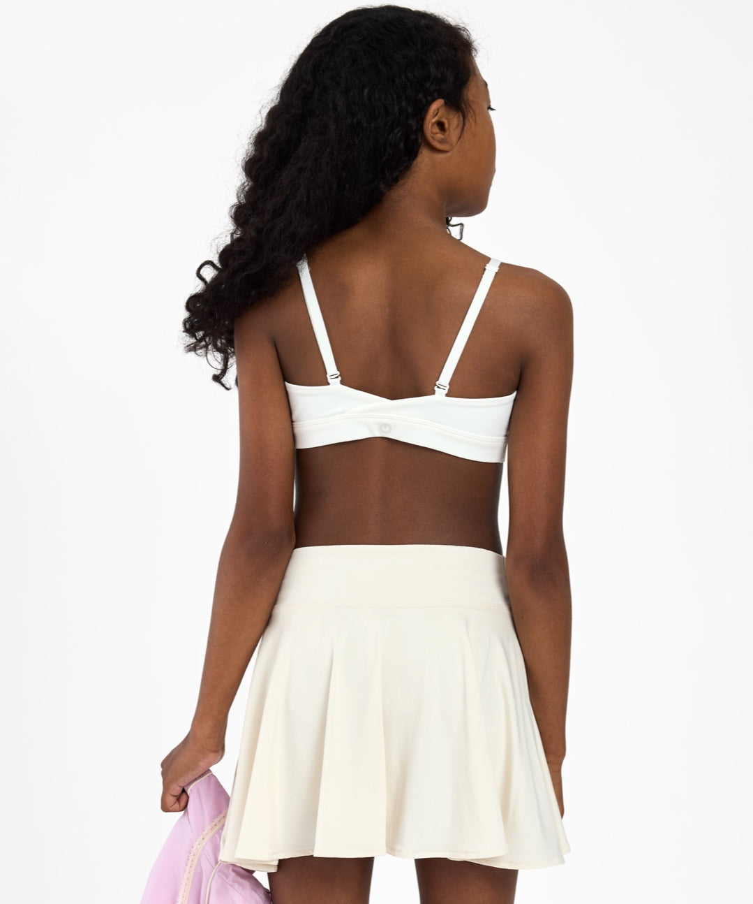 Back view of a girl wearing a white sports bra, holding a pink jacket on a white background.