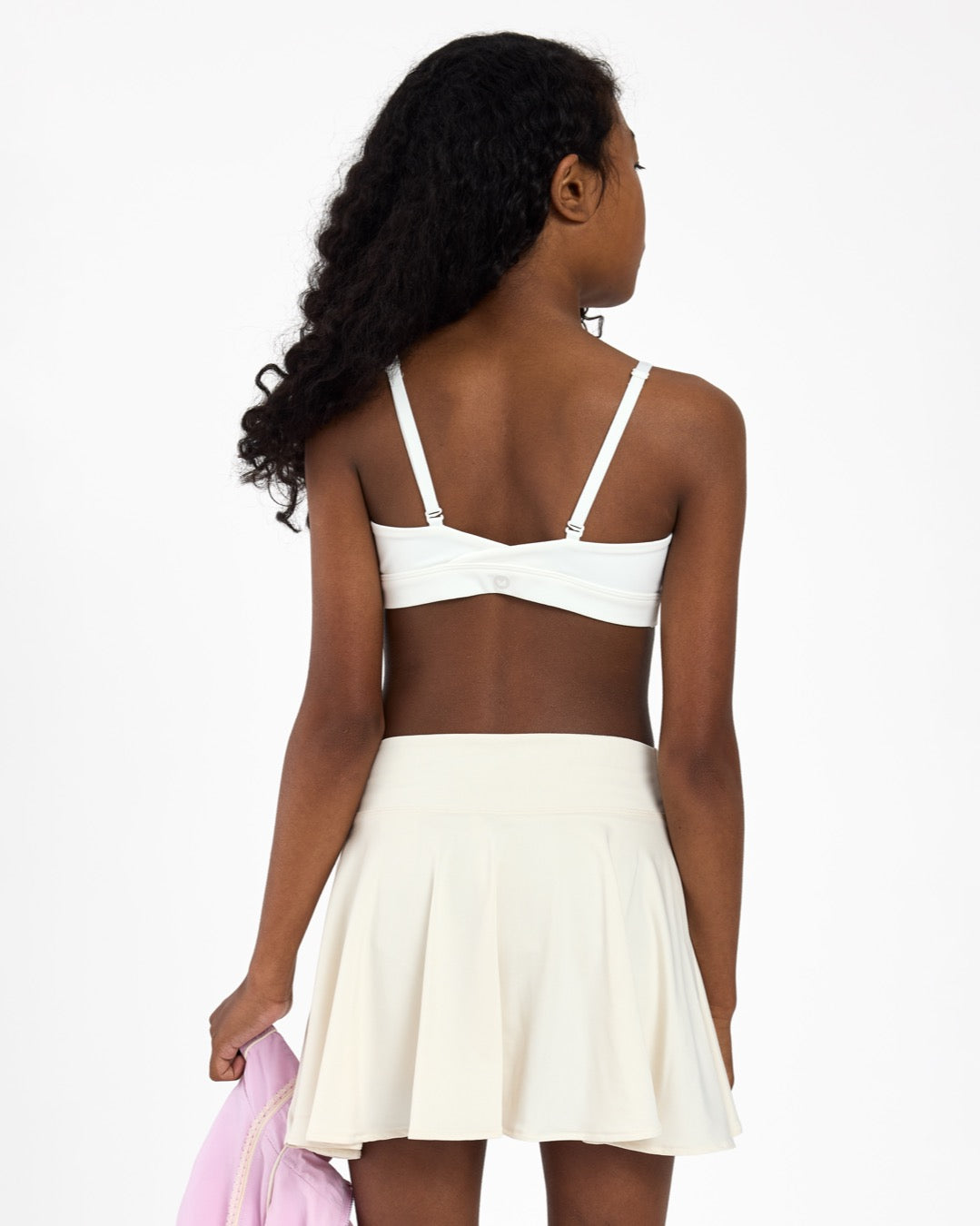 Back view of a girl wearing a white sports bra, holding a pink jacket on a white background.