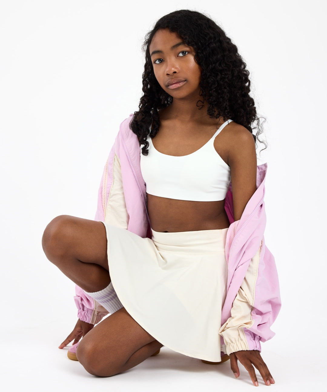 Girl wearing a white first sports bra with a scoop neckline, wearing a pink jacket on a white background.