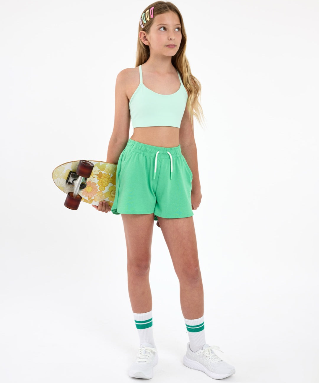 Girl wearing a mint green sports bra and green outfit, holding a skateboard  on a white background.