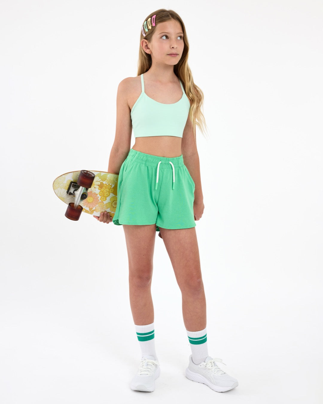 Girl wearing a mint green sports bra and green outfit, holding a skateboard  on a white background.