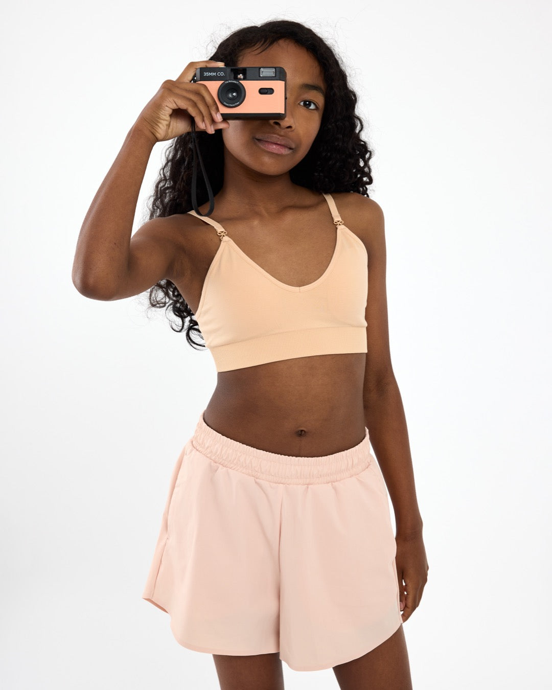 Girl holding a camera wearing a beige crop top training bra with a seamless underband and adjustable straps.