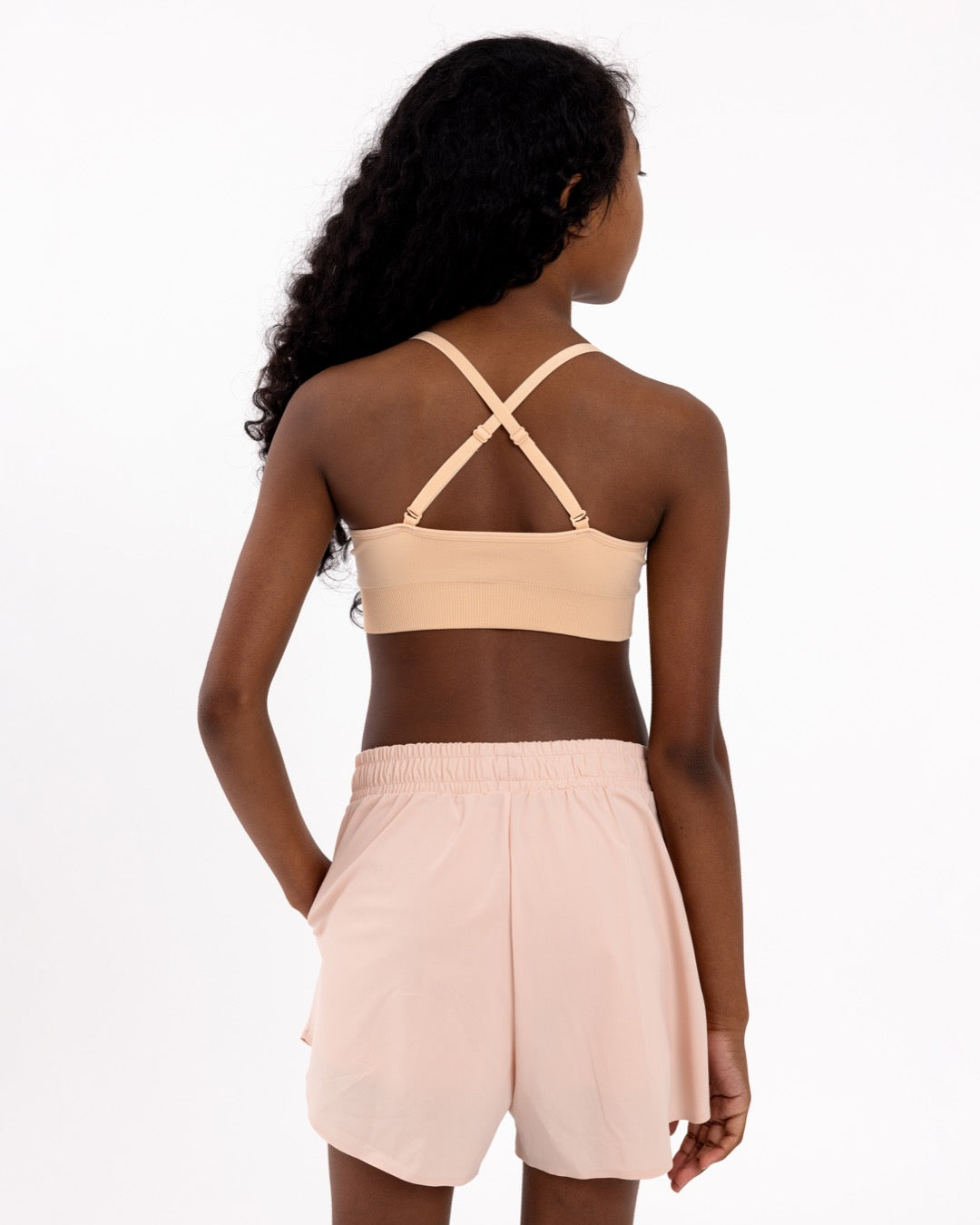 Back view of a girl wearing a beige first training bra showing adjustable crossover straps and seamless underband.