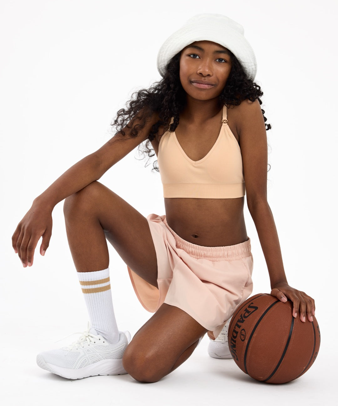 Girl wearing a beige crop top bra with a with adjustable straps, sitting on the floor holding a basketball.