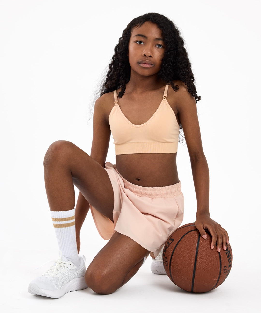 Girl wearing a beige crop top and shorts holding a basketball on a white background.