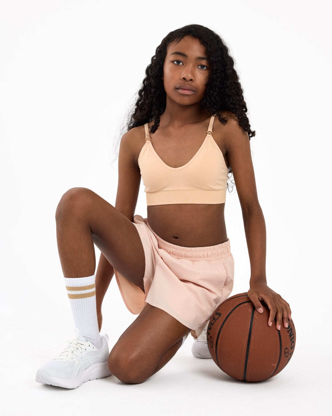 Girl wearing a beige crop top and shorts holding a basketball on a white background.