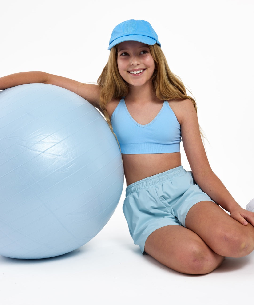 Girl leaning on a Pilates ball wearing a light-blue first training bra with a seamless crop top fit and adjustable straps.