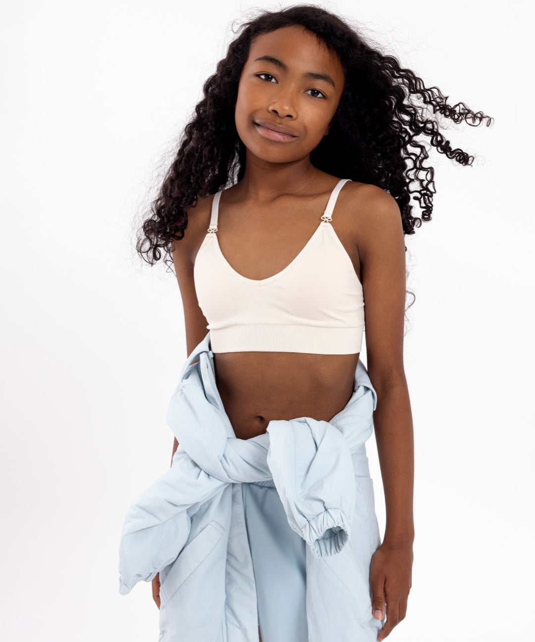 Girl wearing a white crop top with a seamless underband and adjustable straps with bow detail.