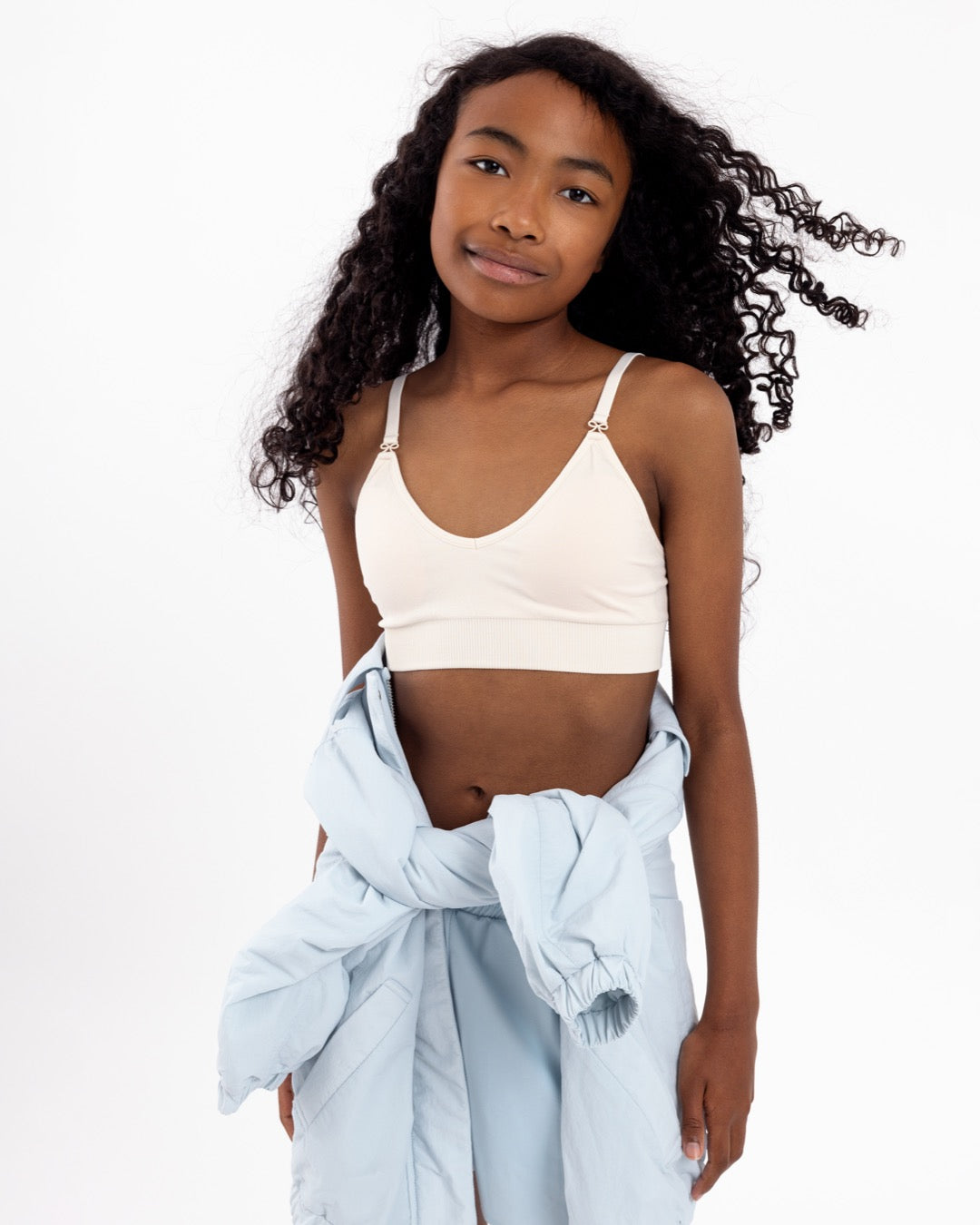 Girl wearing a white crop top with a seamless underband and adjustable straps with bow detail.